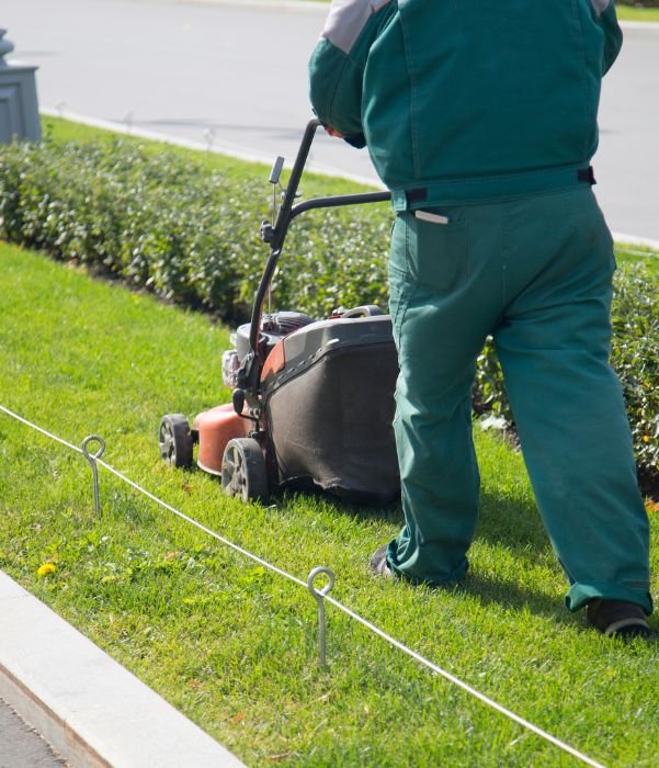 A man wearing green pants and a green shirt is mowing a grassy lawn under clear skies.